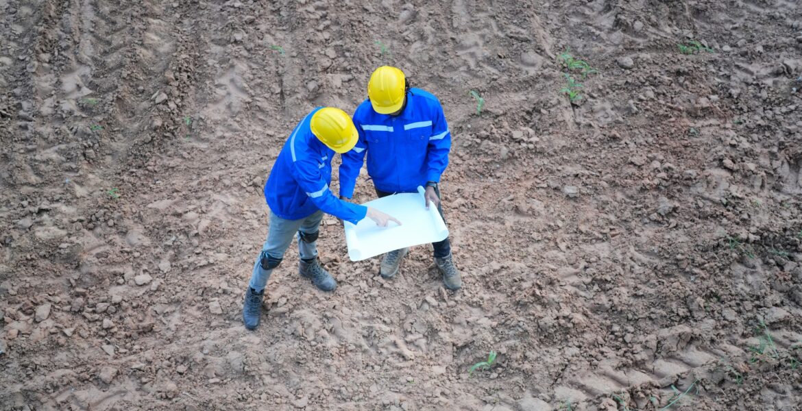 Top view two engineers discussing over blueprint on land at wind turbine site.